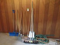 Full view of all garden tools including shovel, rakes, garden hose coiled, and clippers leaning against wood wall