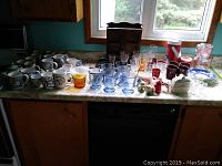 Wide view of collection of coffee mugs, glasses, and some small bowls on kitchen counter under window