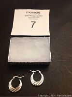 Pair of sterling silver hoop earrings with smooth and ridged design on a dark surface next to a box and auction card.