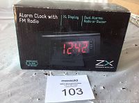 Boxed digital alarm clock showing XL red LED display and key features such as FM radio, dual alarms, and battery backup requirement.