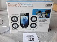 Photo of front of boxed iSound Quad-X portable speaker system showing four speaker drivers and a smartphone dock, USB power cable included.