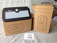 Photo showing one Arilux solar sensor light with LED panel and motion sensor on top of two product boxes with line drawings.