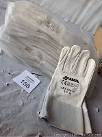 Pair of white AMIA welder gloves with black printed brand and model details on the back. Gloves placed on a flat surface with packaging and lot tag nearby.