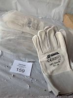 Photo showing a pair of white leather AMIA welding gloves with labeling ART 016/30 TG 10, placed on a plastic surface with a MaxSold lot tag