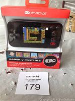 Front view of Gamer V Portable Gaming System in packaging, showing the device screen with gameplay and label indicating 220 games included.