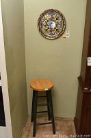 Painted wood stool with natural wood seat and black legs positioned under a wall-mounted decorative plate with floral pattern.