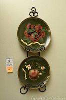 Two 10-inch fruit themed plates displayed vertically on black metal rack hung on wall