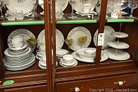 Noritake china set displayed in glass cabinet with multiple plates, cups, and saucers clearly arranged.