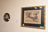 Large Egyptian papyrus artwork in black and gold frame and small oval painting of a woman reading in dark wood frame on wall.