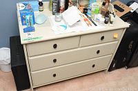 Front view of beige dresser with clutter on top, showing three drawers and black circular knobs.