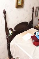 Photo showing one wooden headboard leaning against wall with carved tall posts and curved top rail, covered partially with bed linens and crocheted blanket.