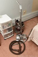 Full view of the Filter Queen vacuum canister with hose and metal wand, placed on carpeted floor next to small plastic drawers and bed.