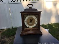 Front view of the Seth Thomas mantel clock, showing wooden frame, Roman numeral clock face with handle on top, placed outdoors on a black surface