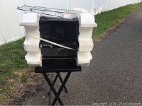 Full view of the black convection oven tightly sealed in white styrofoam packaging, placed on a stand outside.