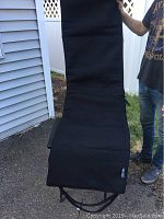 Black full-length massage chair pad with a textured fabric exterior, rectangular shape with slight padding details, being held upright outside near a fence.