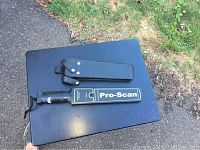 Pro-Scan metal detector placed on a black surface outdoors, showing front side with 'Pro-Scan' label and attached wrist strap.