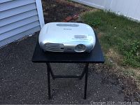 White Epson projector on a black folding table outside, showing side vents and lens.