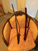 Full view of katana sword resting on a peach upholstered chair alongside its brown wooden scabbard with red cord detail.