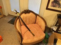 Ceremonial saber fully sheathed in its black and brass scabbard laid on a pink upholstered chair.