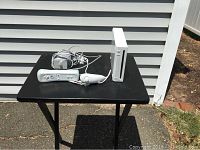 White Nintendo Wii console and wired Wii Remote controller with Nunchuk on small black table outside.