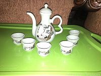 White porcelain Japanese tea set with one teapot and six cups with dragon and floral decoration placed on a green plastic surface.