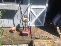 Front view of Craftsman mini tiller/cultivator near shed, showing red body, handles, and fuel tank.