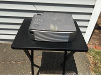 Photo of the Mitsubishi RS 232c projector placed on a black folding table, showing top view with cosmetic marks and wear.