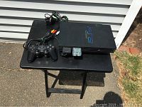 Black PlayStation 2 console on black table outdoors showing controller and AV cables with red, white, and yellow connectors.