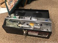 Opened black metal toolbox containing a MasterGrip HVLP gravity feed spray gun, several metal cups, and accessories arranged inside.