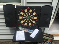 Front view of black wooden dartboard cabinet with electronic dartboard inside, including packaged darts and white cloths below.
