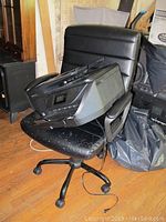 Black adjustable office chair with HP OfficeJet 6958 printer placed on the seat, showing wear and dust on both items.