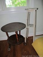 Photo showing round wooden table and wooden plant stand next to it against white wall and window