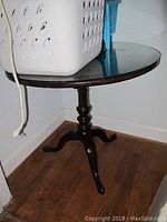 Photo of round wooden pedestal table top with a white laundry basket and blue chair partially visible on top, showing surface wear and scratches.