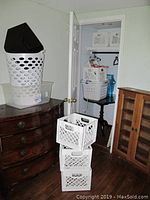 Photo of two white laundry baskets stacked with black plastic basket inside, clear plastic storage bin below, and three white plastic milk crates stacked beside furniture in room.