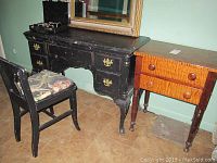 Photo shows black wooden desk with brass hardware and drawers next to wooden chair with floral padded seat and warm brown end table with two drawers and a golden framed mirror on wall.