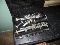 Disassembled clarinet inside a black soft-lined case resting on a dark table surface. Parts include the upper and lower body, barrel, bell, and mouthpiece with some cork missing on joints.