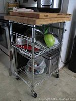 Side angle showing the rolling kitchen cart with a wood block top, wire shelves with pots and bowls, and kitchen appliances on and inside the cart.