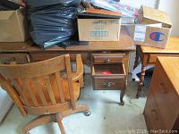 Desk with drawers and wood finish, chair with slatted backrest and casters, side table visible in background with turned legs.