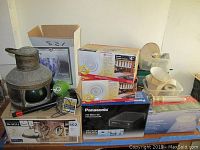 Overview of lot showing stacked boxes of various lighting kits, a Panasonic DVD burner box and a vintage metal lamp with green globe on wooden table.