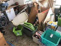 Several green and clear plastic storage bins, a white plastic shower seat, and assorted household and storage items piled in a garage or storage area.