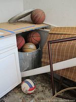 Photo showing multiple basketballs including an NBA ball stored in a metal trash can and on a shelf above near unassembled basketball hoop parts.