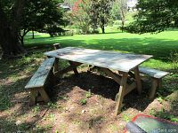 Photo showing outdoor wooden picnic table with two benches in green lawn setting, weathered paint visible on table top and benches.