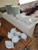 Wide view showing stacked salad plates and saucers along the edge of a counter beside cups arranged in front.