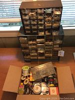 Full view of two stacked metal hardware cabinets with multiple drawers and an open cardboard box filled with various hardware items on a table in front of a window.