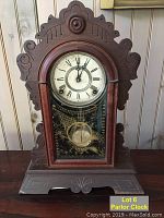 Front view of late 19th century wooden parlor clock with carved wood details, Roman numeral clock face and glass front door.