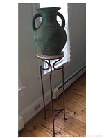 Full view of green ceramic vase sitting on round sandstone top supported by metal stand in corner of room