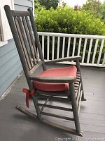 Side view of the weathered grey wooden porch rocking chair on a porch with a tied red cushion on the seat.