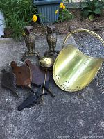 Overall collection including 3 bellows, brass wood holder, owl andirons, and boot remover on concrete ground near garden.