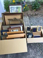 Two boxes filled with approximately three dozen picture frames of various sizes and styles from small to large, seen outdoors with gravel and plants in background.