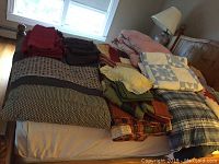 Overall photo of quilts placed on a bed showcasing multiple folded quilts and bedding sets in various colors and patterns including red, brown, blue, yellow, and multicolor quilts.